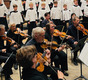 Eine Gruppe von Musikern mit Violinen sitzt im Vordergrund, während ein Kinder- und Jugendchor in weißen Gewändern im Hintergrund steht. Die Musiker konzentrieren sich auf ihre Instrumente, während der Chor singt. Die Szene spielt in einer festlichen Umgebung.