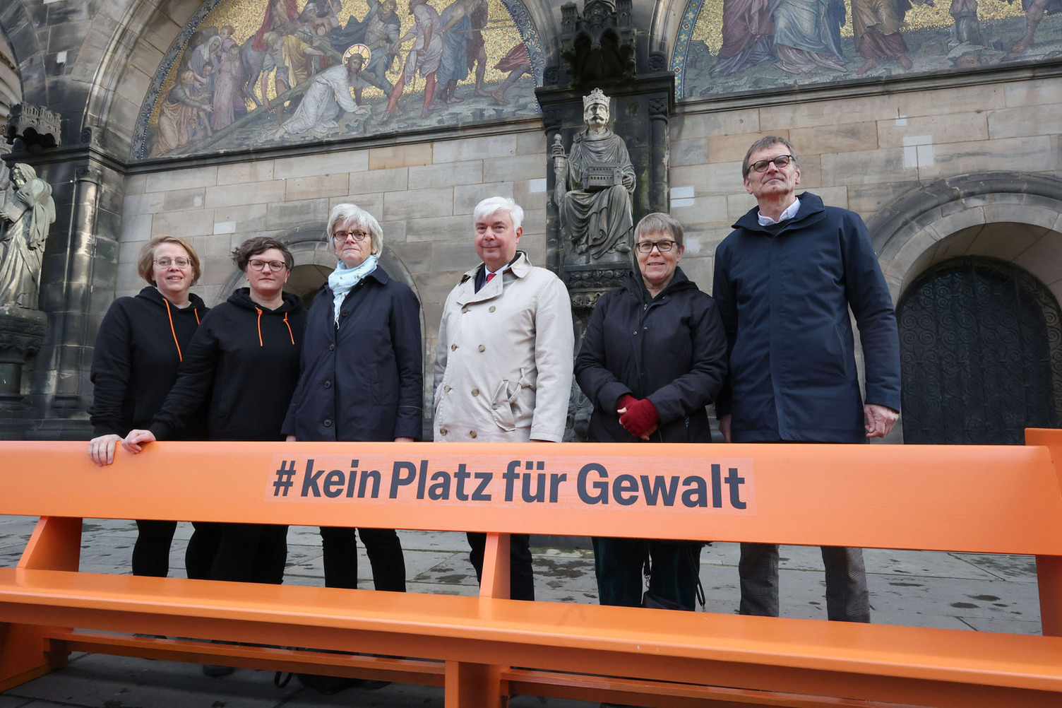 Auf dem Foto ist eine orangefarbene Bank mit der Aufschrift #KeinPlatzFürGewalt zu sehen. Hinter der Band stehe Präsidentin Edda Bosse, Schriftführer Pastor Bernd Kuschnerus und Jutta Schmidt. Auf der Bank sitzen  Pastorin Ragna Miller, Nancy Janz und Dompastor Henner Flügger.   