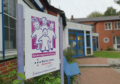 Ein Schild mit der Aufschrift „Kindertageseinrichtung“ und dem Logo der Martin Luther Gemeinde ist vor einem Gebäude angebracht. Im Hintergrund sind Teile des Gebäudes und eine Sitzbank sichtbar.