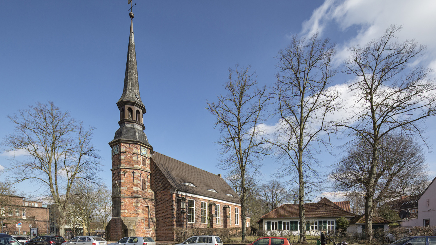 Ein historisches Kirchengebäude mit einem hohen, spitzen Turm und einer Uhr an der Fassade. Umgeben von kahlen Bäumen und geparkten Autos, unter einem klaren blauen Himmel mit einigen Wolken.