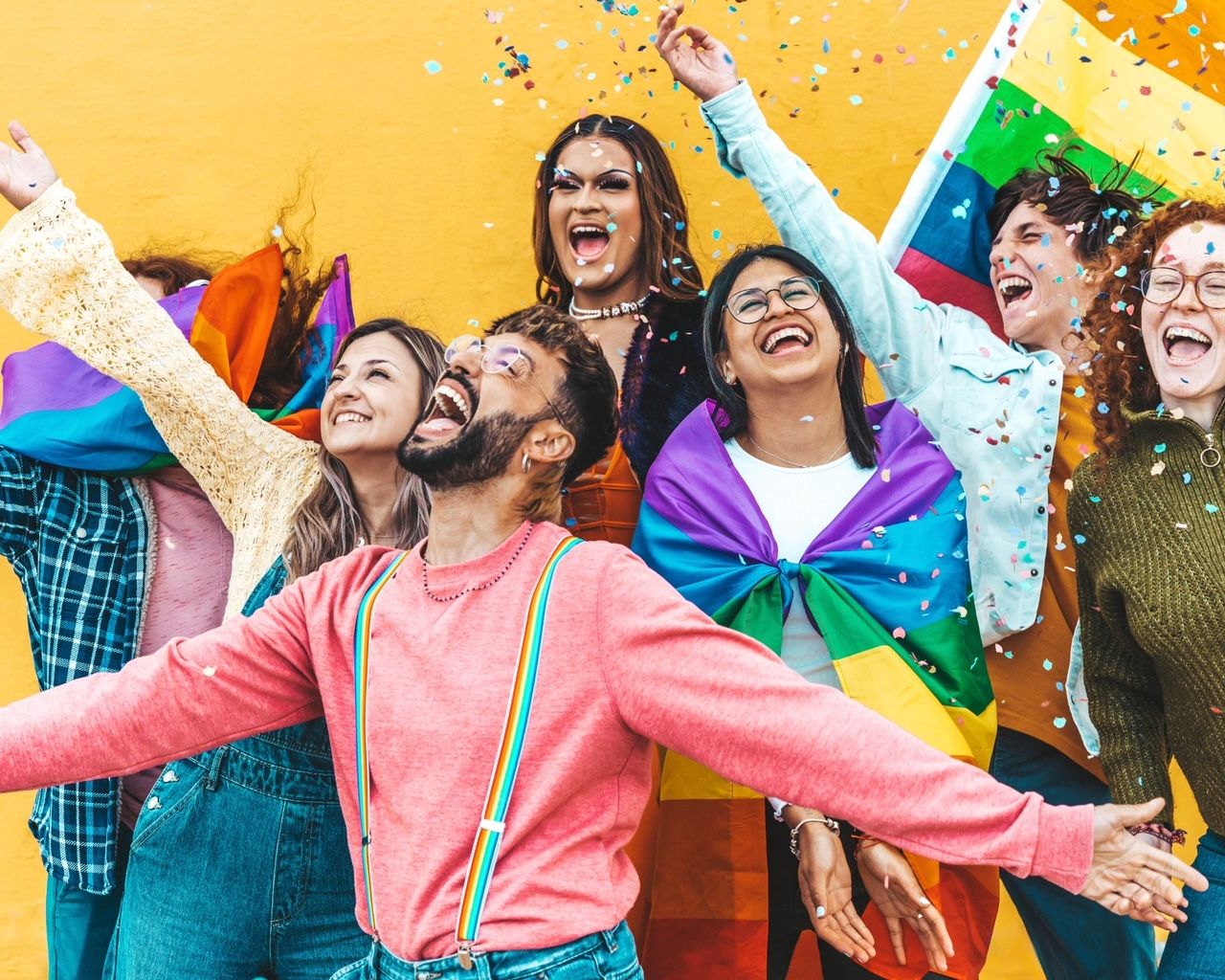 Eine Gruppe von lachenden, gut gelaunten Menschen ist zu sehen. Sie sehen ganz verschieden aus, tragen bunte Kleidung und Regenbogenflaggen bei sich. Über ihnen schwebt buntes Konfetti durch die Luft.