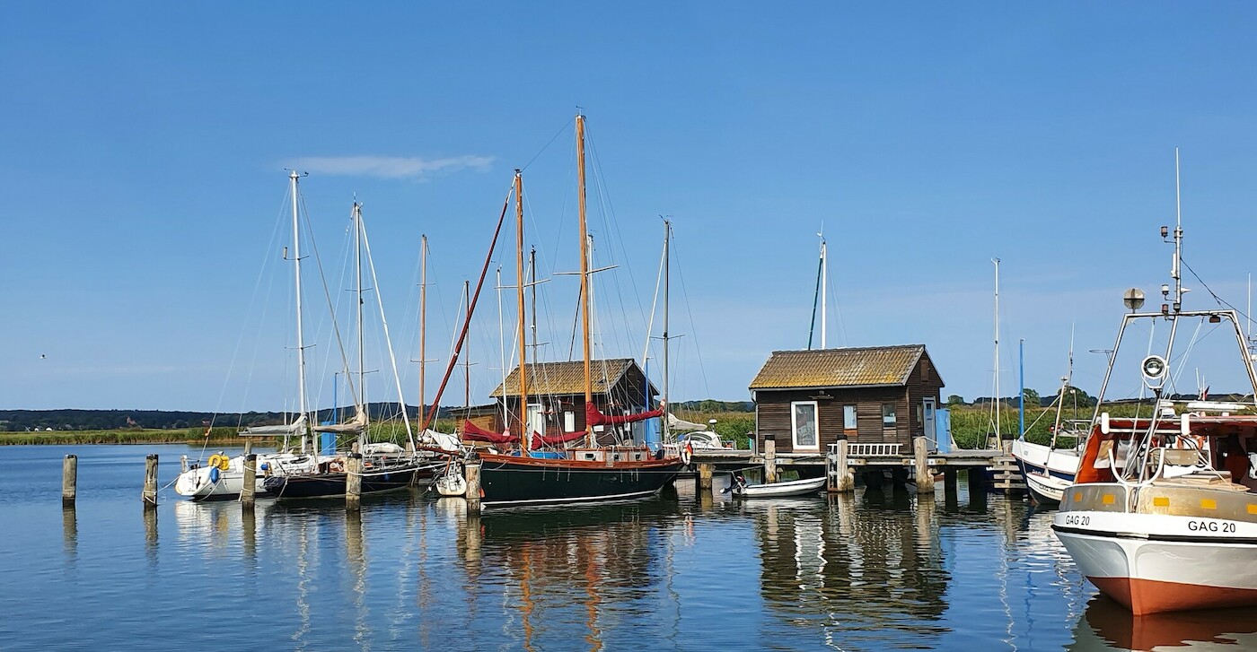 Zu sehen ist ein Hafen mit 2 Hütten, an dem Segelschiffe anliegen. Das Wasser ist ruhig und der Himmel ist wolkenfrei blau.