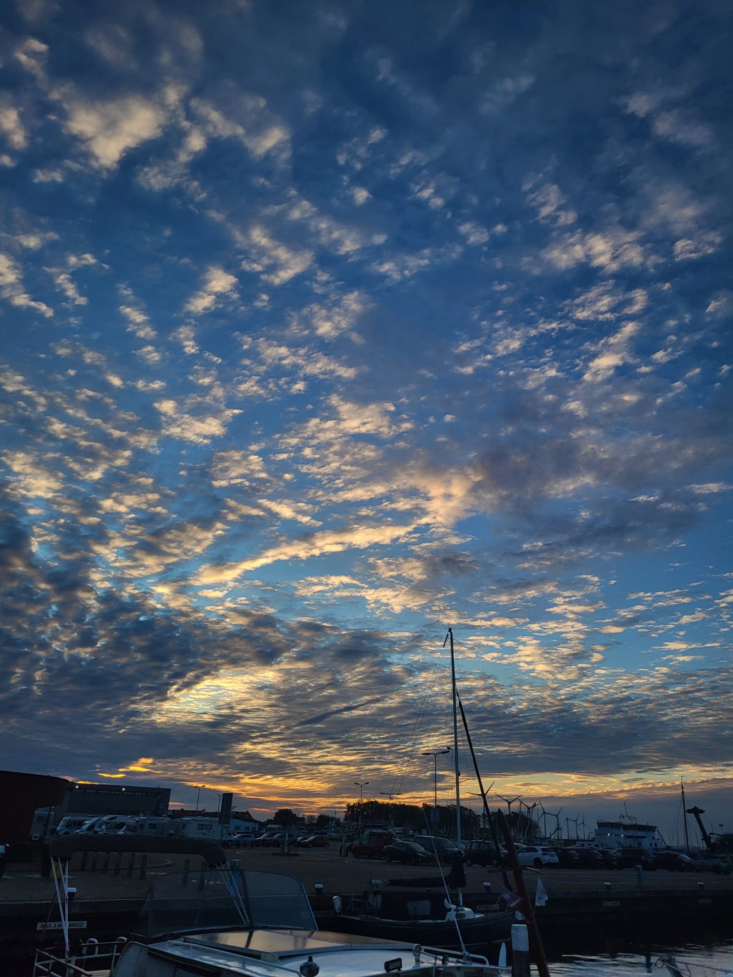Urk Sonnenaufgang Der Himmel zeigt ein beeindruckendes Farbspiel bei Sonnenaufgang, mit zahlreichen Wolken in Blau- und Gelbtönen. Im Vordergrund sind Teile eines Hafens und Boote zu erkennen, während die Silhouetten von Gebäuden und einem Parkplatz im Hintergrund verschwommen sind.