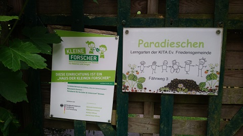 Zwei Schilder an einem Zaun: Links steht "Kleine Forscher" mit Informationen über das Haus. Rechts zeigt das Schild "Paradieschen", Lern- und Gartenbereich der evangelischen Friedensgemeinde. Es gibt eine Adresse: Fährweg 3.