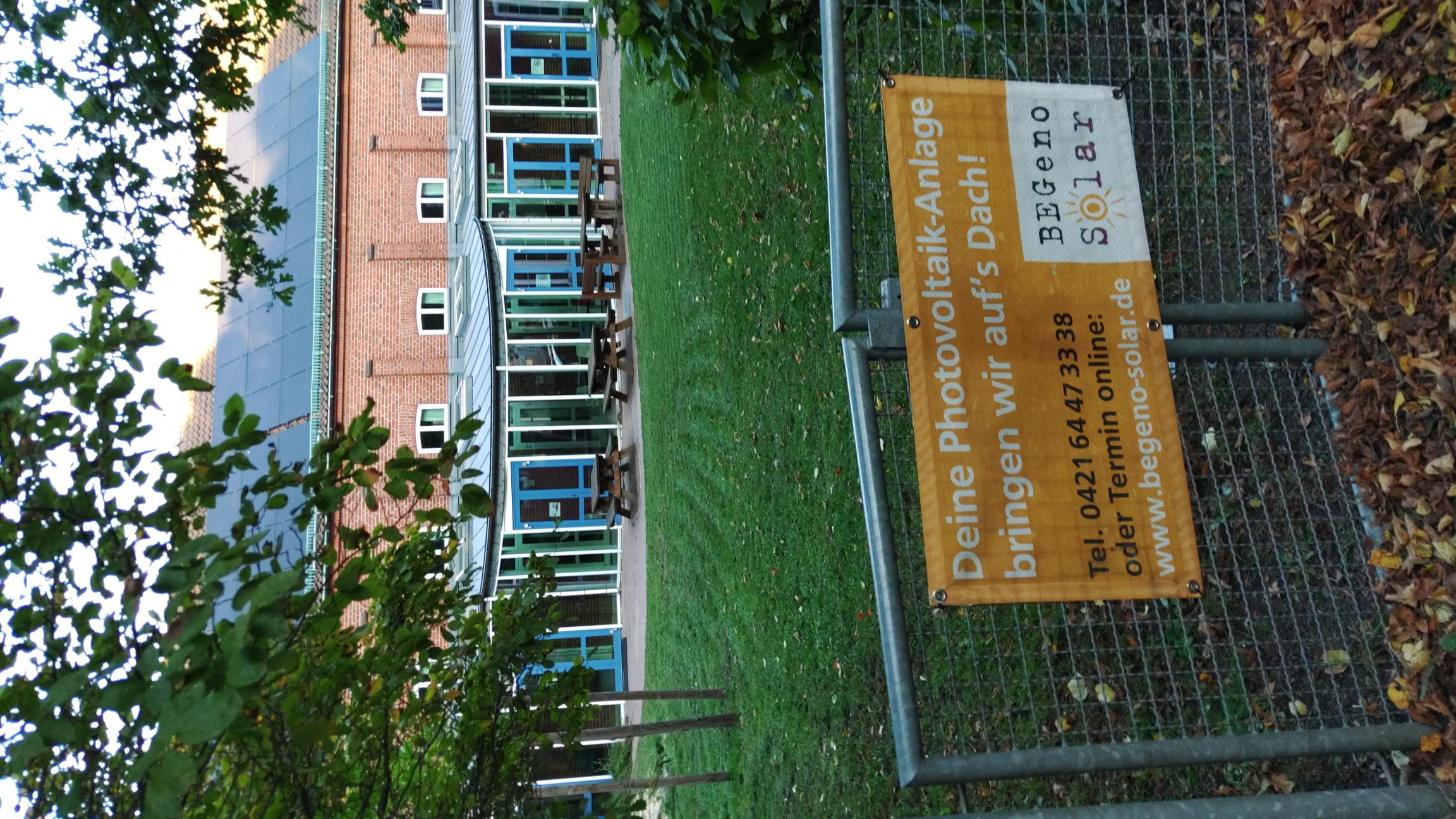 Der eingezäunte Außenbereich der Martin-Luther-Gemeinde in Findorff. Am Zaun hängt ein großes Schild, dass die Photovoltaik-Anlage der Gemeinde beschreibt, es enthält Kontaktinformationen. Im Hintergrund ist ein modernes Gebäude mit großen Fenstern und einer Photovoltaikanlage auf dem Dach zu sehen.