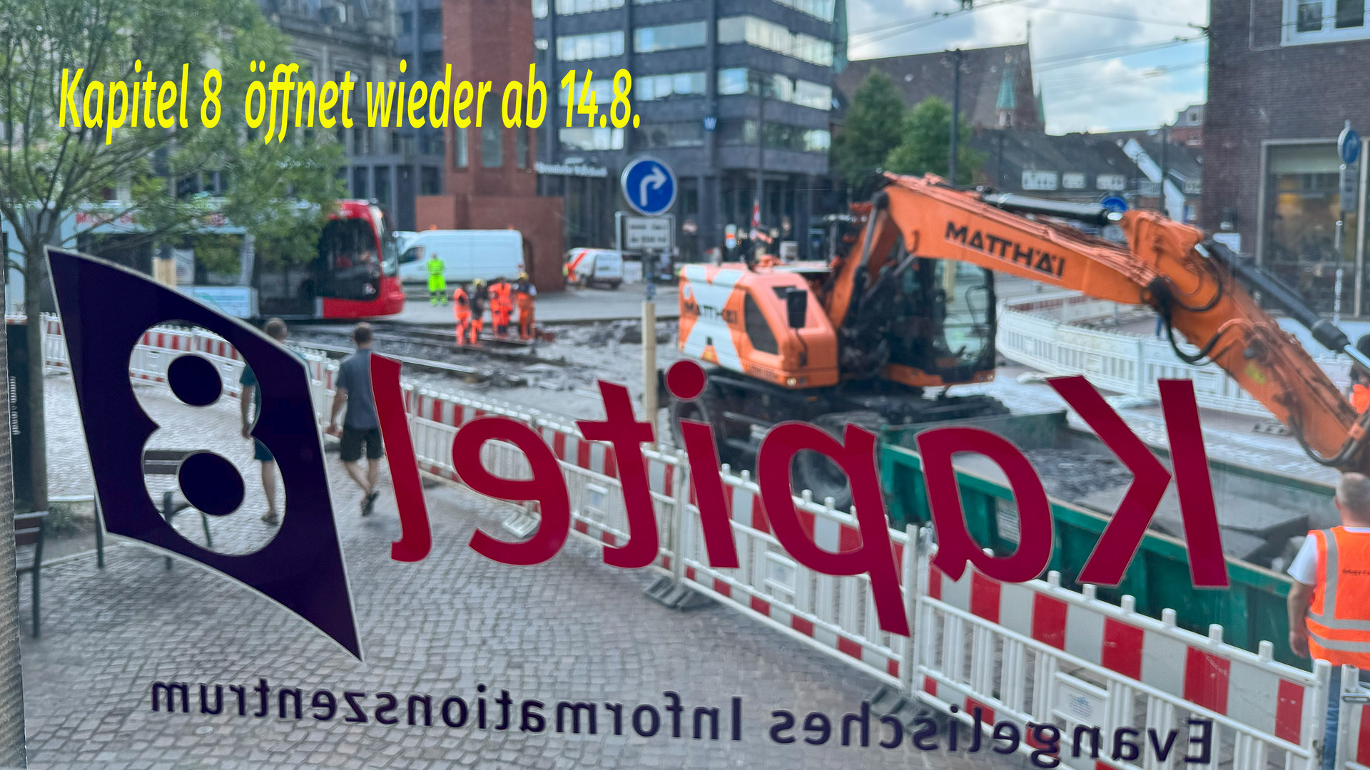 Auf dem Bild ist ein Fenster mit der Aufschrift „Kapitel 8 öffnet wieder ab 14.8.“ zu sehen. Im Hintergrund sind Bauarbeiten sichtbar, mit einem großen Bagger und Mitarbeitern in Warnkleidung, die an einer Baustelle arbeiten.
