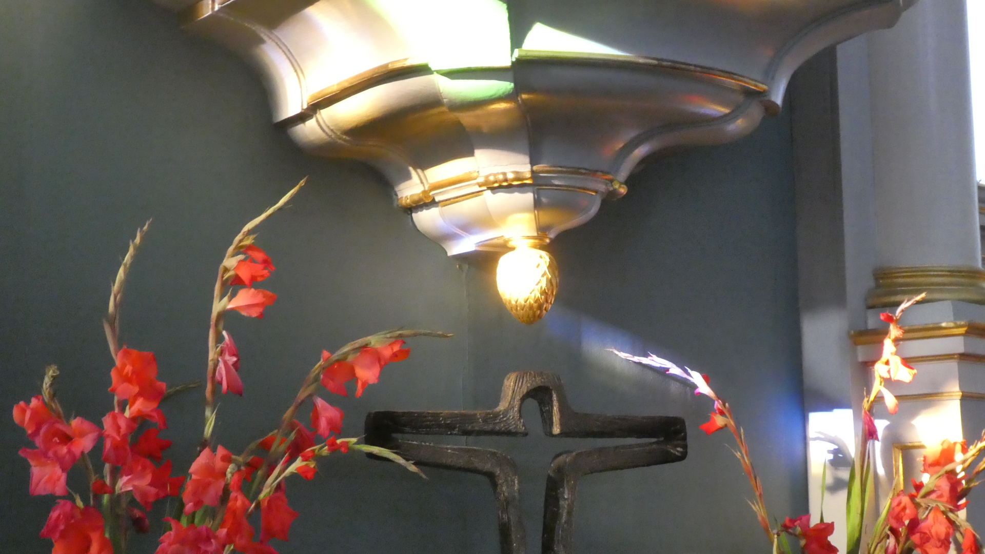 Eine Kreuzdarstellung aus Holz ist vor einem dunklen Hintergrund zu sehen, flankiert von roten Blumen. Oberhalb des Kreuzes befindet sich eine goldene Lampe, die sanftes Licht auf die Szene wirft, wodurch eine ruhige, feierliche Atmosphäre entsteht.