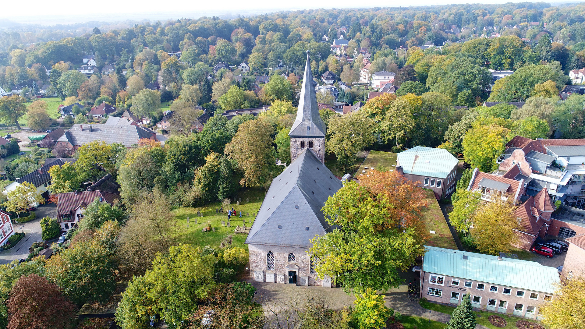 Luftaufnahme einer Kirche mit spitzem Turm, umgeben von bunter Herbstvegetation. Im Vordergrund sind Gräber und eine Wiese sichtbar. Im Hintergrund sind Wohngebäude und Bäume, die die ländliche Umgebung in lebhaften Farben präsentieren.
