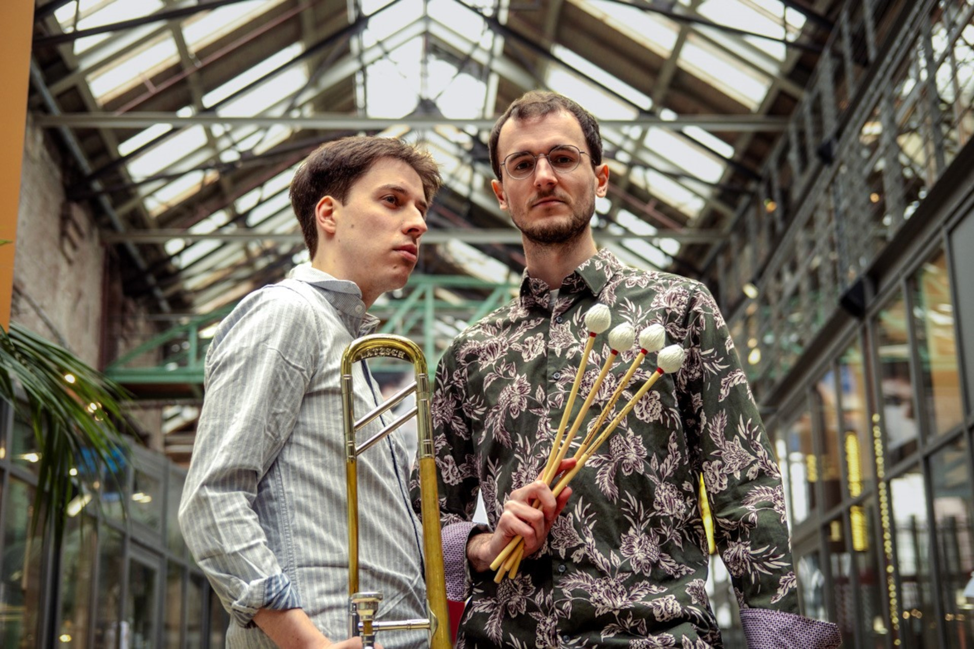 Zwei Musiker stehen in einem lichtdurchfluteten Raum. Der erste hält eine Posaune, während der zweite vier Schlägel in der Hand hält. Beide blicken ernst in die Kamera und sind musikalisch gekleidet. Ihre Umgebung hat ein modernes, industrielles Ambiente.