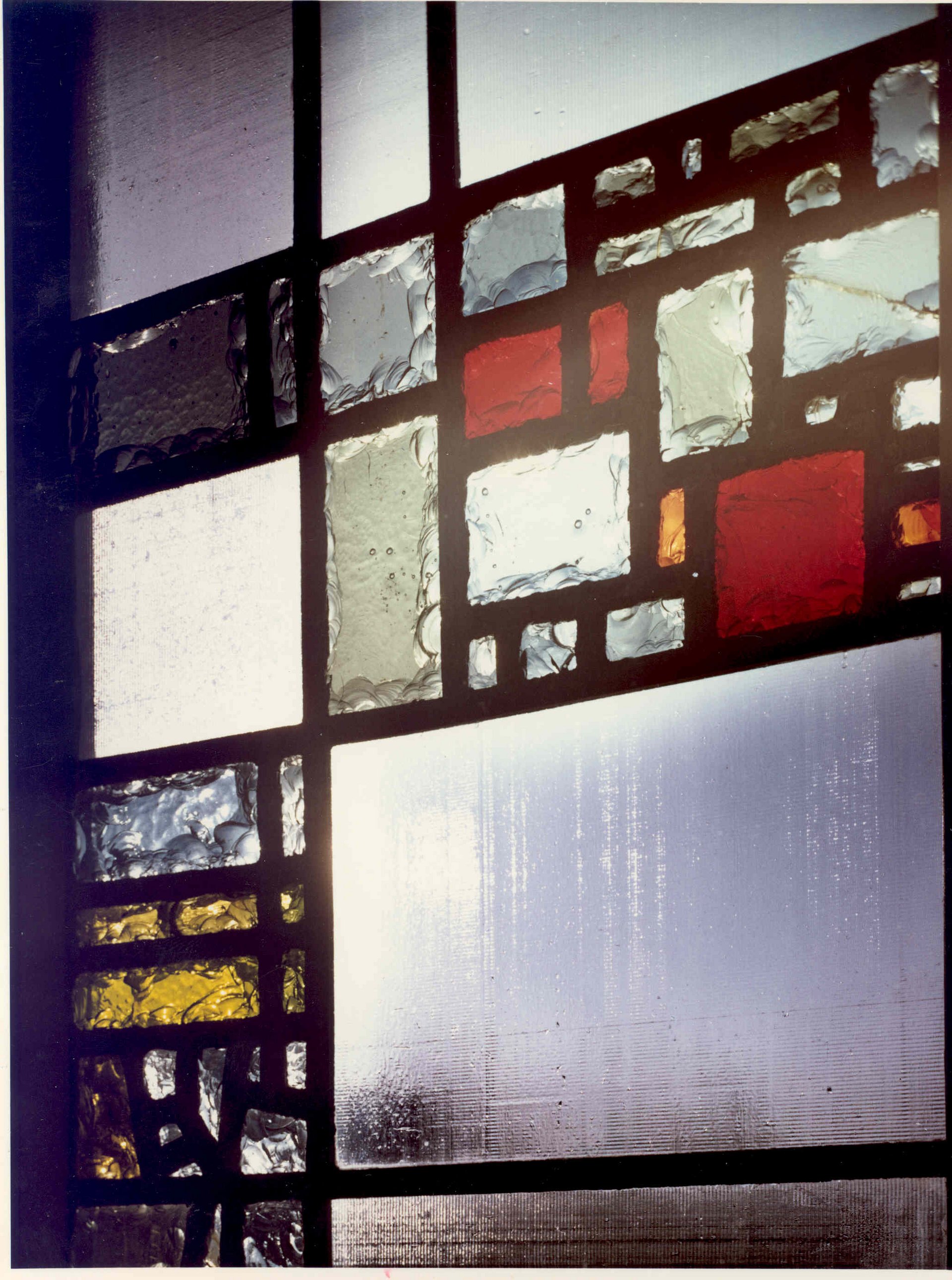 Ein farbiges Glasfenster mit geometrischem Muster, das verschiedene Farbtöne wie Rot, Grün, Gelb und Blau enthält. Die einzelnen Glasstücke sind von schwarzem Glasrahmen umgeben und weisen unterschiedliche Texturen auf, wodurch Licht durchgelassen wird und ein spielerisches Lichtspiel entsteht.