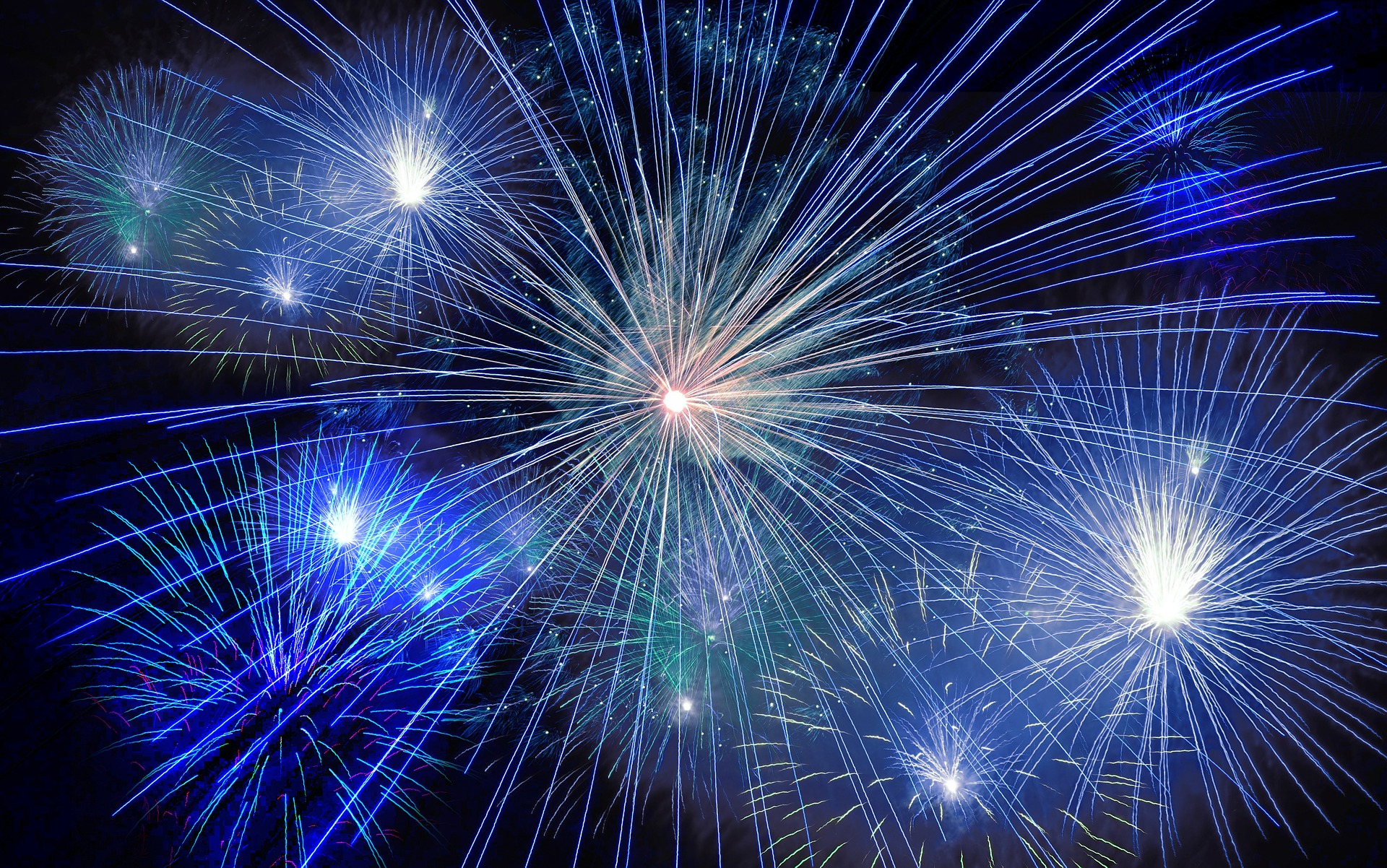 Das Bild zeigt ein spektakuläres Feuerwerk mit bunten Lichtstrahlen in Blau, Weiß und Grün, die in verschiedenen Richtungen strahlen. Der Hintergrund ist dunkel, wodurch die Lichter besonders intensiv leuchten und eine festliche Stimmung erzeugen.